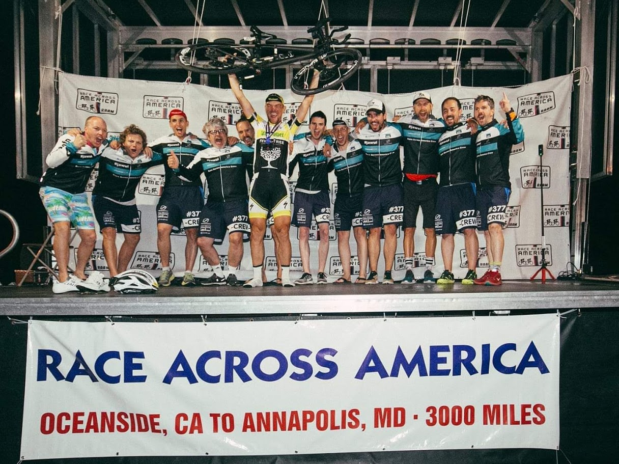 Bild von der Siegerehrung beim Race Across America 2019. Christoph Strasser stemmt sein Fahrrad in die Höhe. Sein Team rund um ihn jubelt mit.